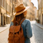 Woman with tan hat and brown backpack walking down a sunlit cobblestone street.